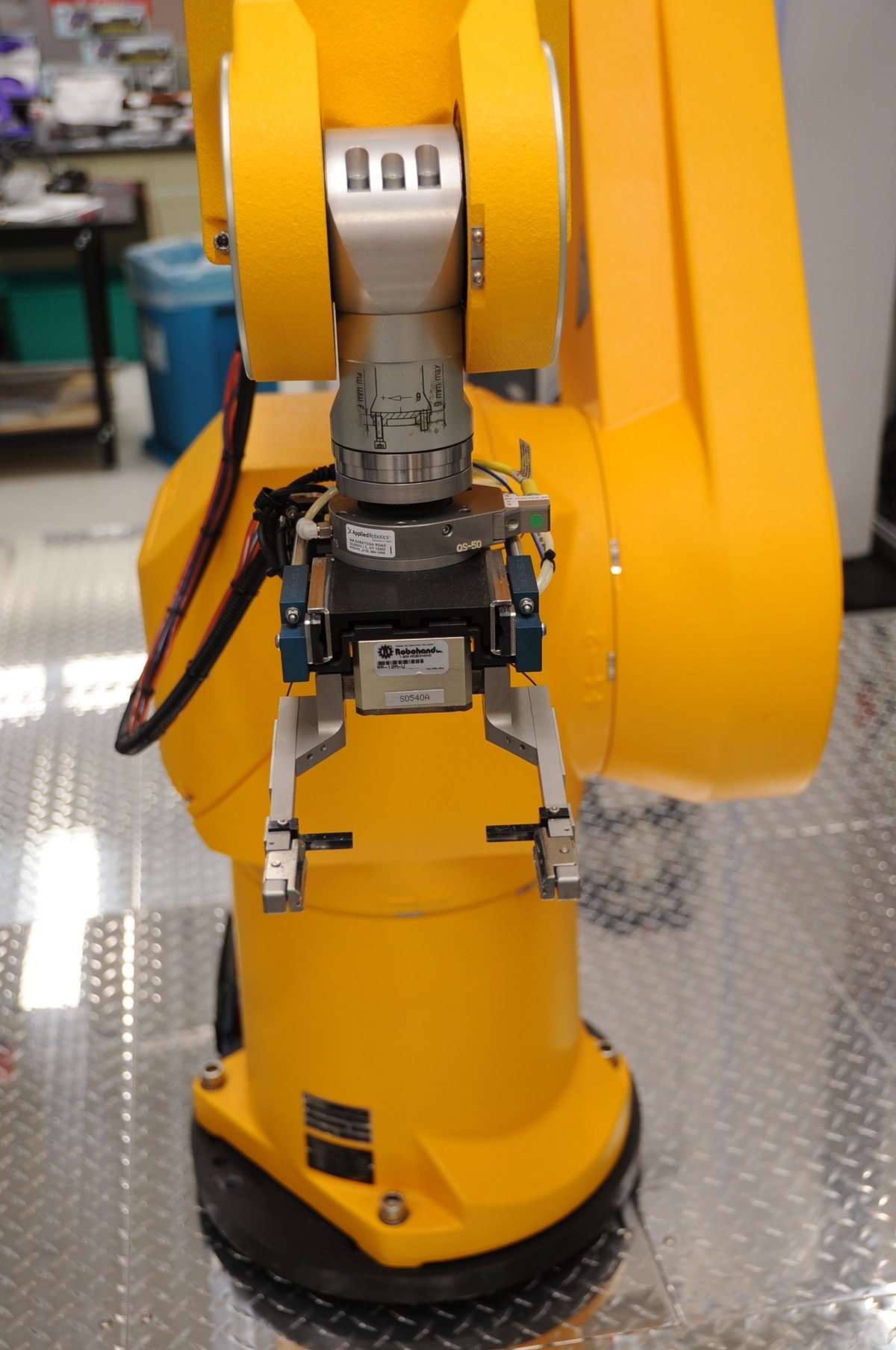 A yellow robotic arm with a gripper standing on a metal floor beside screening equipment